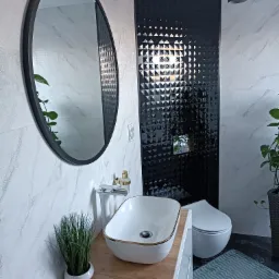 Nowoczesna łazienka w Twardowicach z umywalką nablatową z elementami złota, okrągłym lustrem, czarną ścianą z trójwymiarowych płytek i marmurowymi płytkami na pozostałych ścianach.