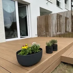 Jasny taras z desek na dwóch poziomach, donice z kwiatami, bambusowa osłona, biały budynek z dużym oknem przesuwnym. Słoneczny dzień.