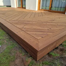 Nowoczesny taras z drewna o unikalnym wzorze ułożenia desek, przylegający do budynku z dużymi oknami, otoczony zielenią.