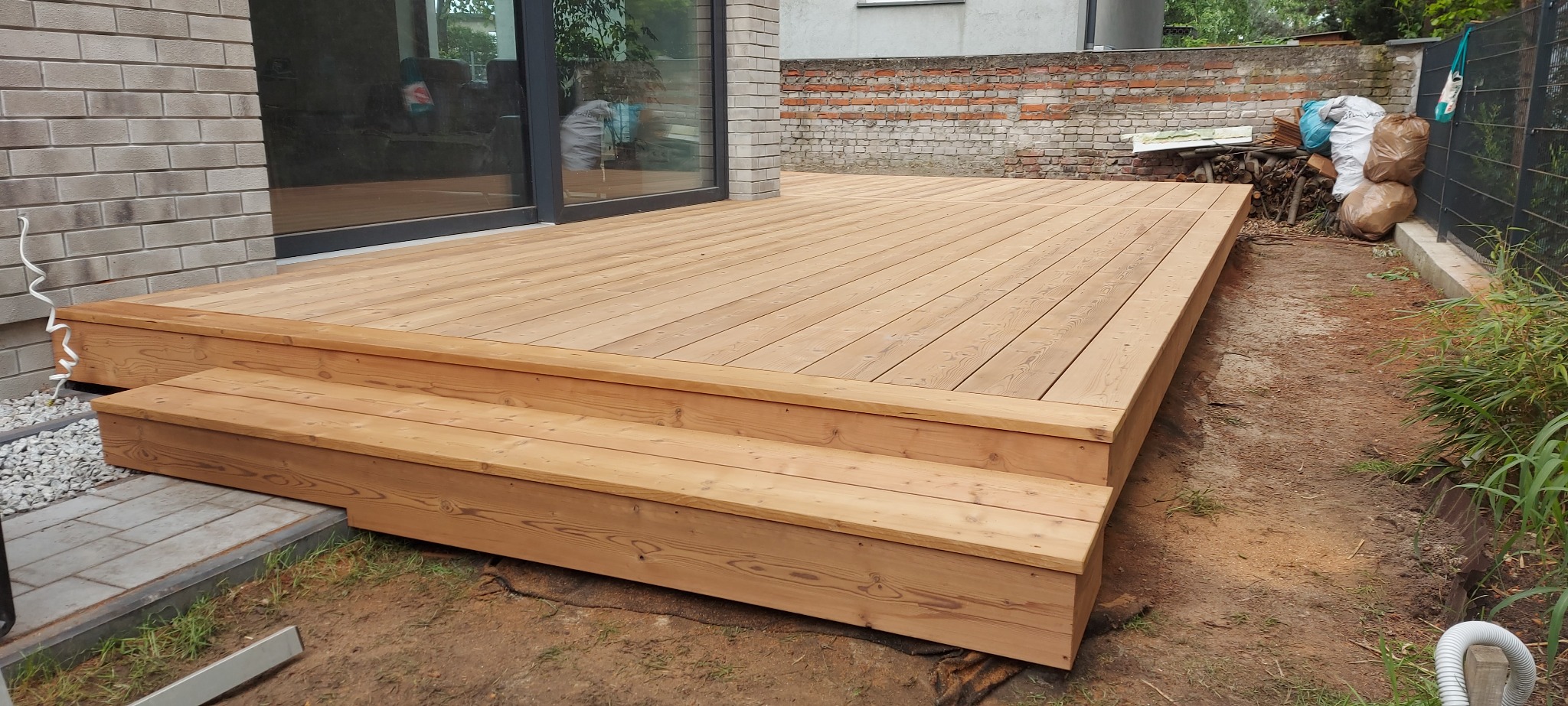 Jasny, drewniany taras z dwustopniowymi schodami, przylegający do nowoczesnego domu z cegły, z widocznymi elementami ogrodu i muru w tle.