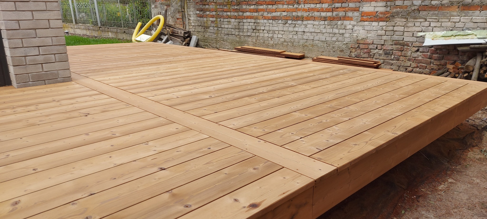 Jasny, drewniany taras z widoczną strukturą drewna, z fragmentem muru z cegły w tle. Elementy konstrukcyjne tworzą regularny wzór na powierzchni.