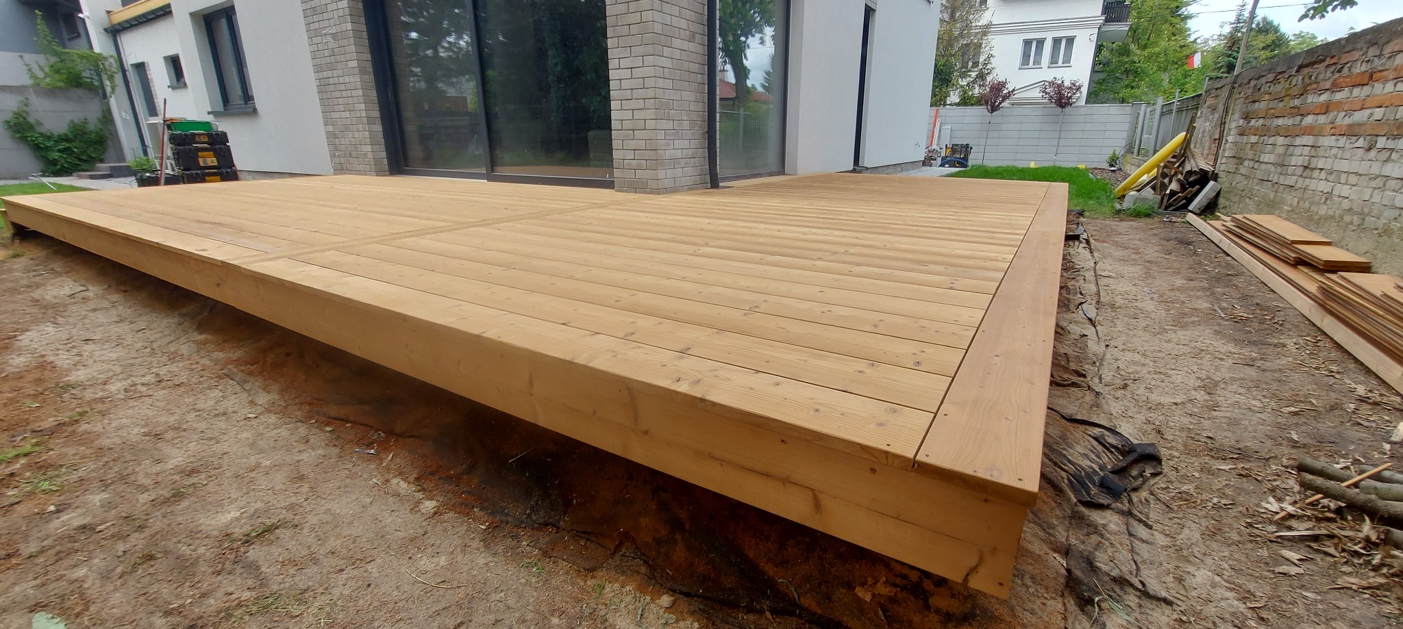 Jasny, drewniany taras przy nowoczesnym domu z ceglaną elewacją. Widoczne deski tarasowe i elementy konstrukcyjne. Częściowo ułożone deski obok.