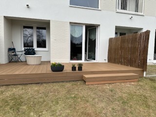 Jasny taras z kompozytu z niskimi schodkami i ażurową przesłoną. Donice z kwiatami, krzesło i stolik na tle jasnego budynku mieszkalnego.