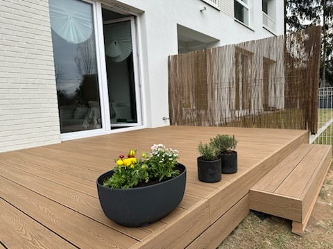 Jasny taras z desek na dwóch poziomach, donice z kwiatami, bambusowa osłona, biały budynek z dużym oknem przesuwnym. Słoneczny dzień.