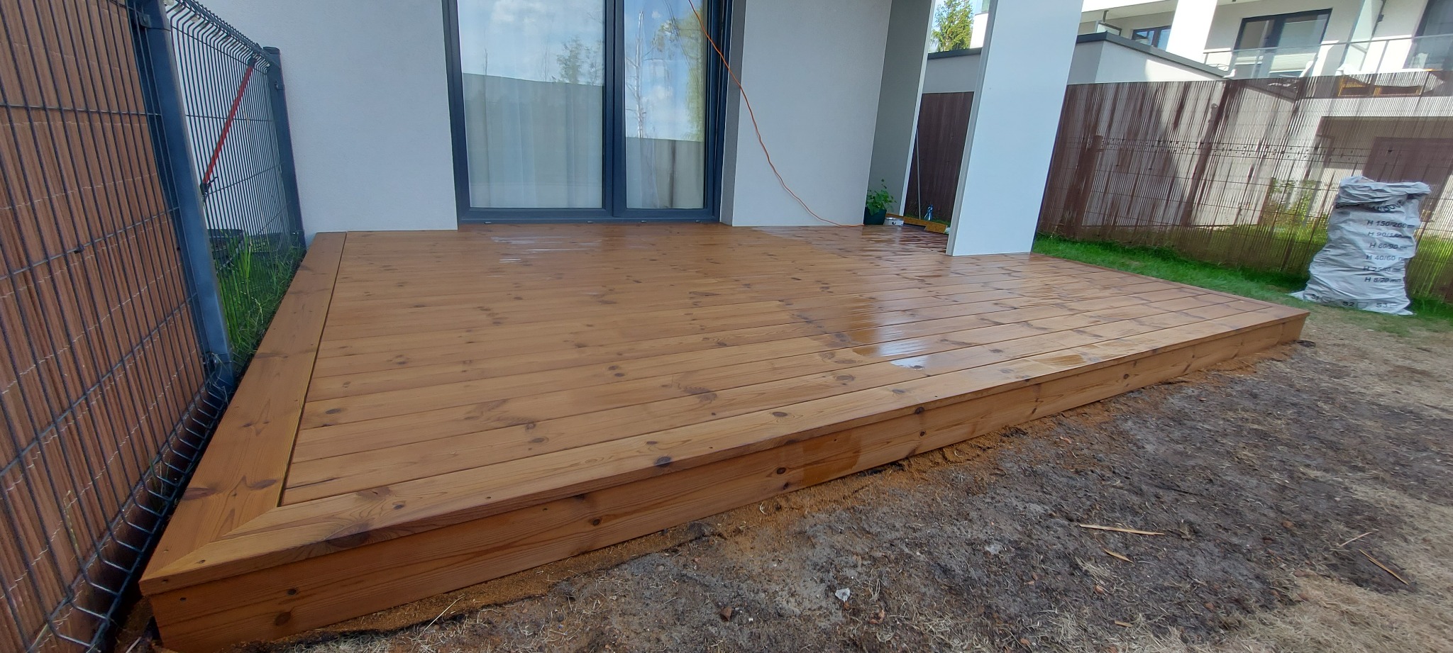 Świeżo zaimpregnowany, drewniany taras przydomowy w odcieniach brązu, z widocznymi słojami drewna, sąsiadujący z ogrodzeniem i budynkiem.