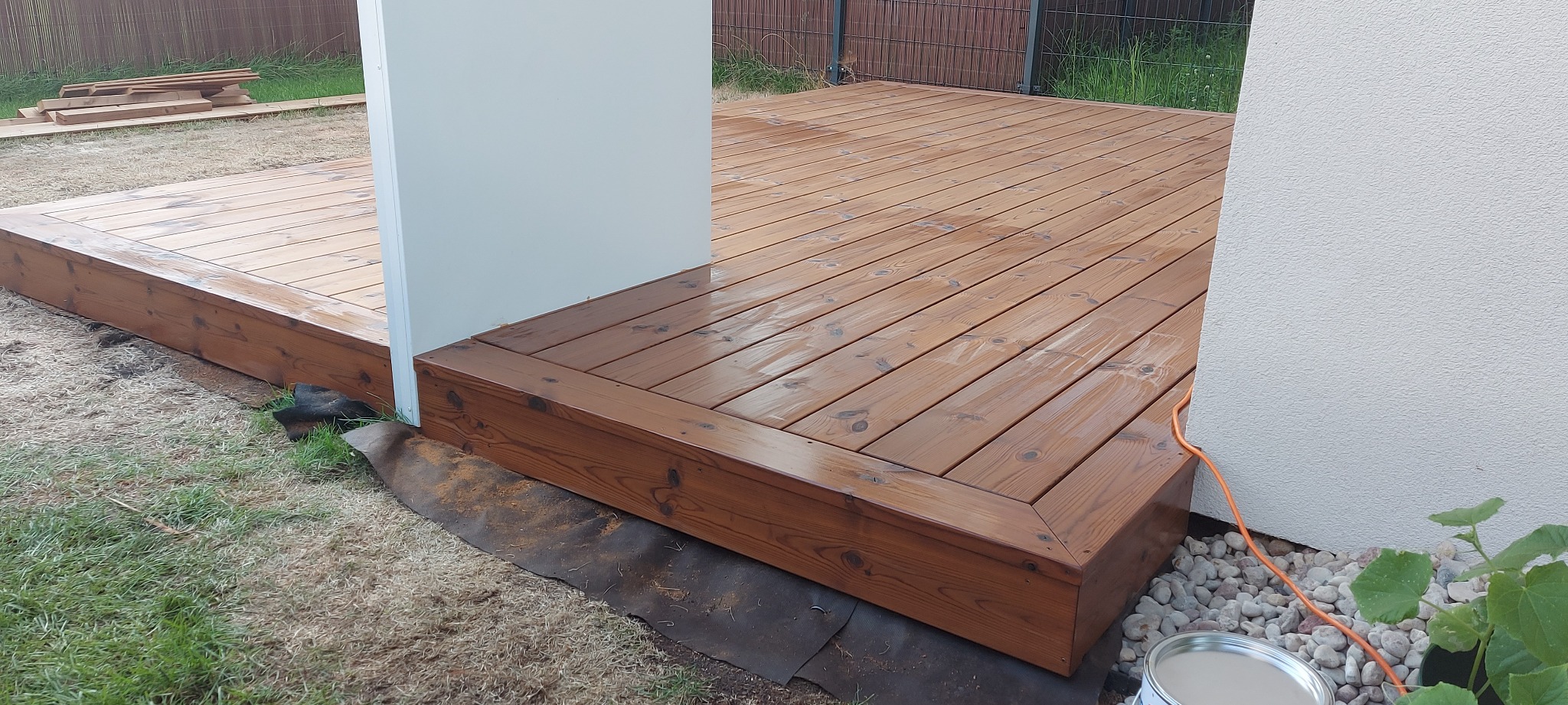 Świeżo polakierowany drewniany taras przydomowy z widoczną puszką farby i detalami wykończenia, brązowe deski, jasna elewacja budynku, kamienie ozdobne.