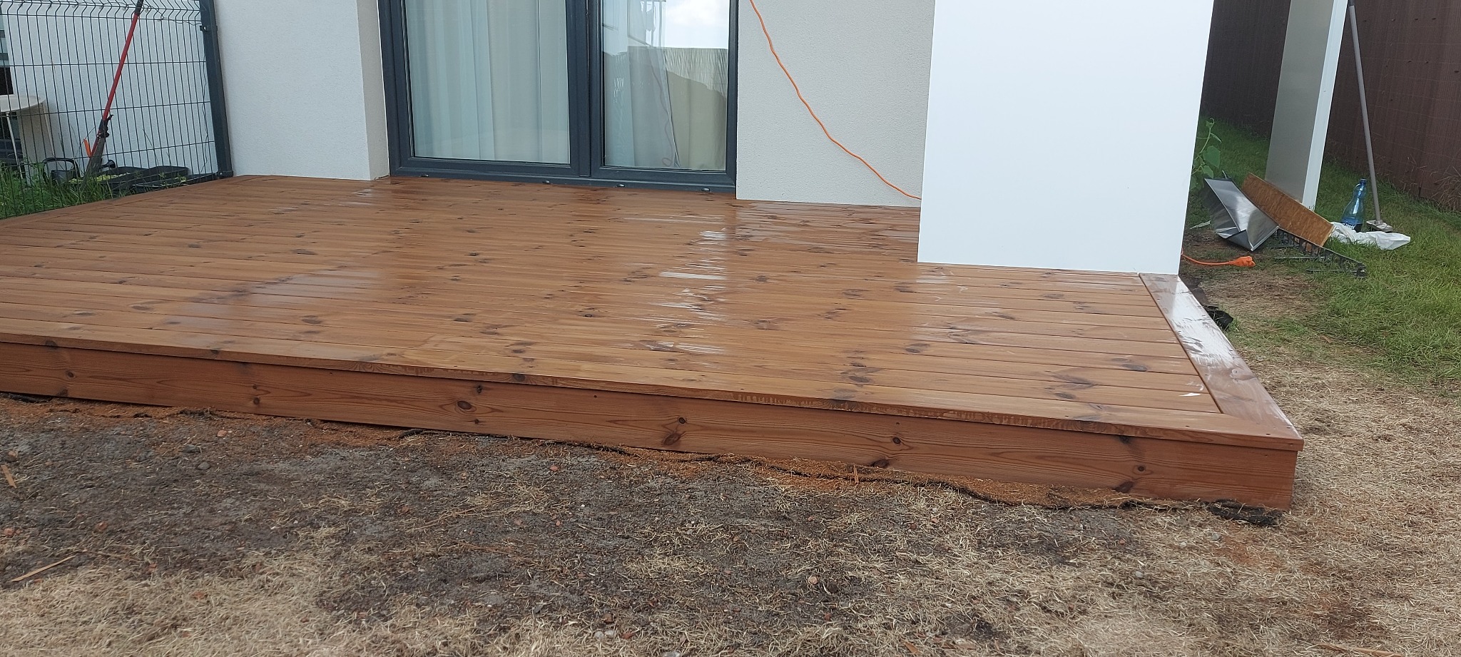 Świeżo zaimpregnowany drewniany taras przydomowy z widoczną fakturą desek i elementami wykończeniowymi, na tle białej elewacji budynku z szarymi oknami.