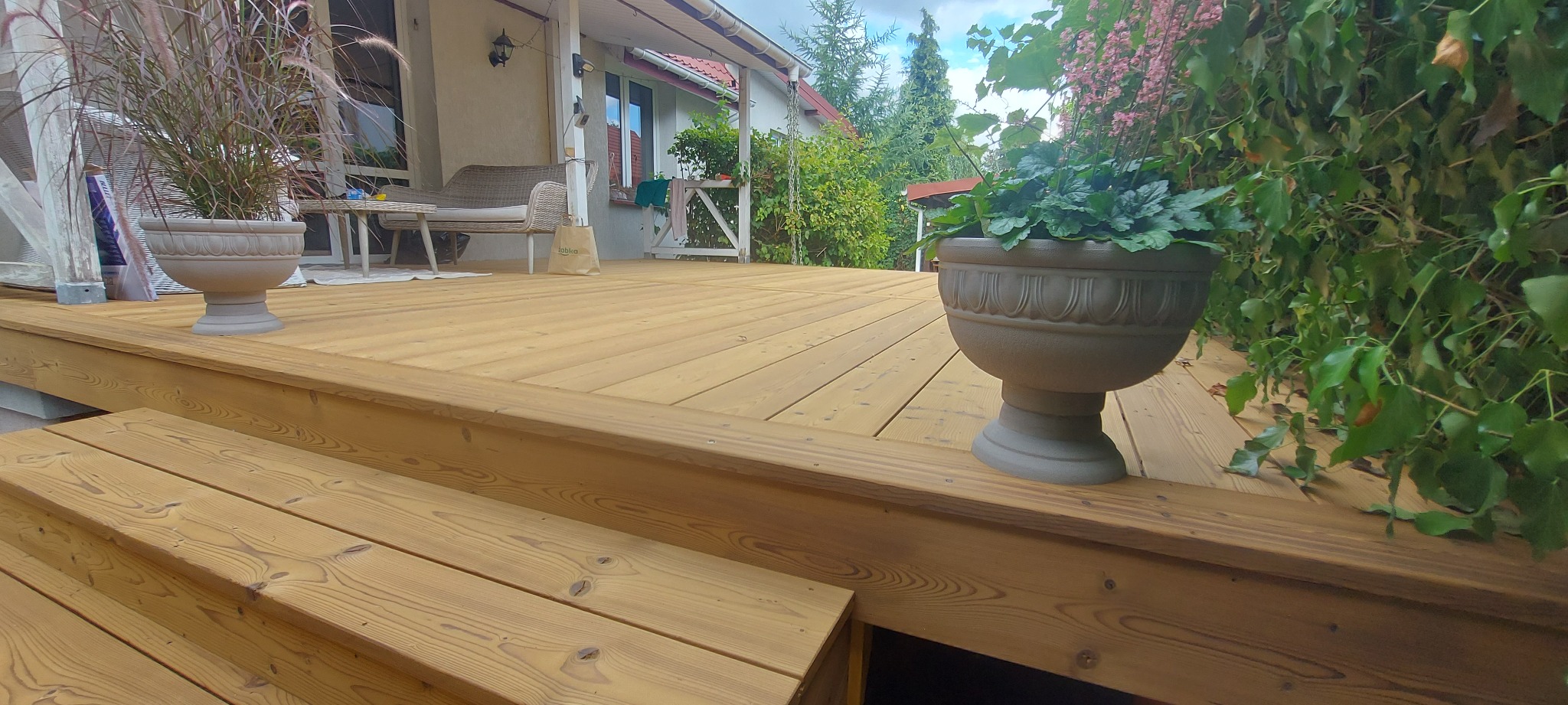 Jasny, drewniany taras z donicami i schodami prowadzącymi do domu. Widoczne meble ogrodowe w tle. Naturalne oświetlenie.