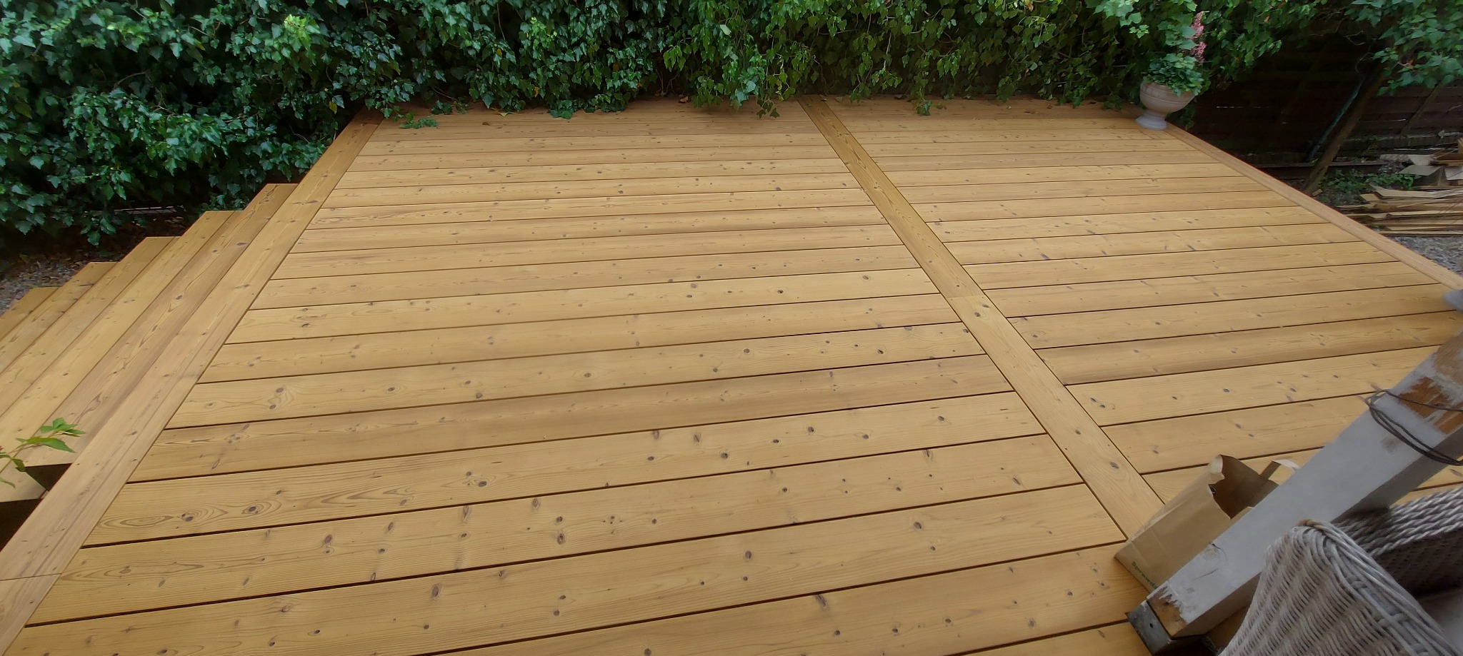 Jasny, drewniany taras z desek, widok z góry, otoczony zielenią. Schody z desek po lewej stronie. Donica z kwiatami w tle.