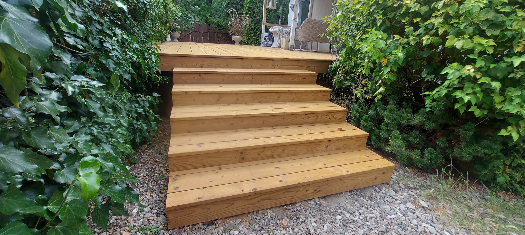 Naturalny drewniany taras z schodami, otoczony bujną zielenią. Widok na fragment tarasu z meblami ogrodowymi w tle.