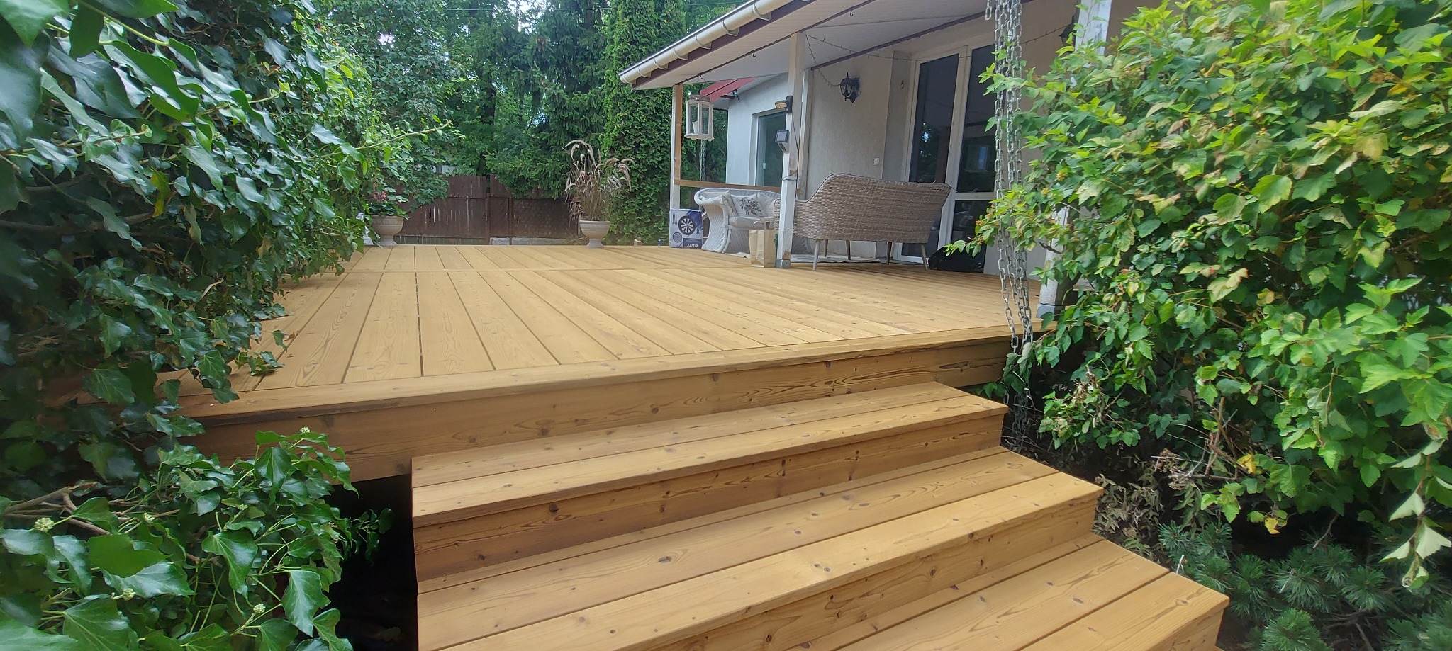 Jasny, drewniany taras z trzema stopniami schodów, otoczony zielenią. W tle biały budynek z wiklinową sofą. Widok z bliska, naturalne oświetlenie.