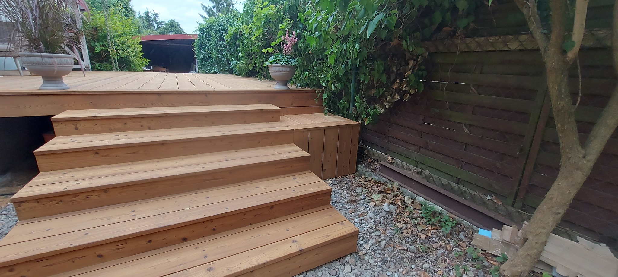 Drewniany taras z schodami, otoczony zielenią i ogrodzeniem. Widoczne donice z kwiatami. Naturalne oświetlenie, ciepłe barwy drewna. Ujęcie z dołu, podkreślające strukturę drewna.