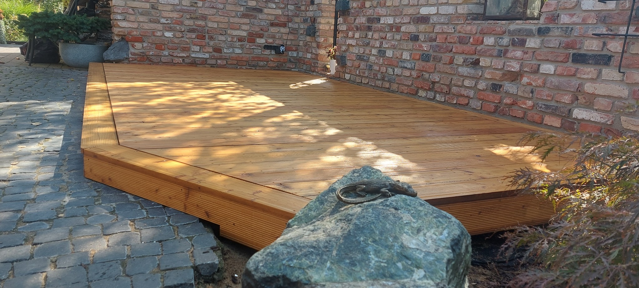 Jasny, drewniany taras przylegający do ceglanego muru. Uwagę przykuwa dekoracja w postaci kamienia z figurką jaszczurki na pierwszym planie. Zewnętrzne oświetlenie.