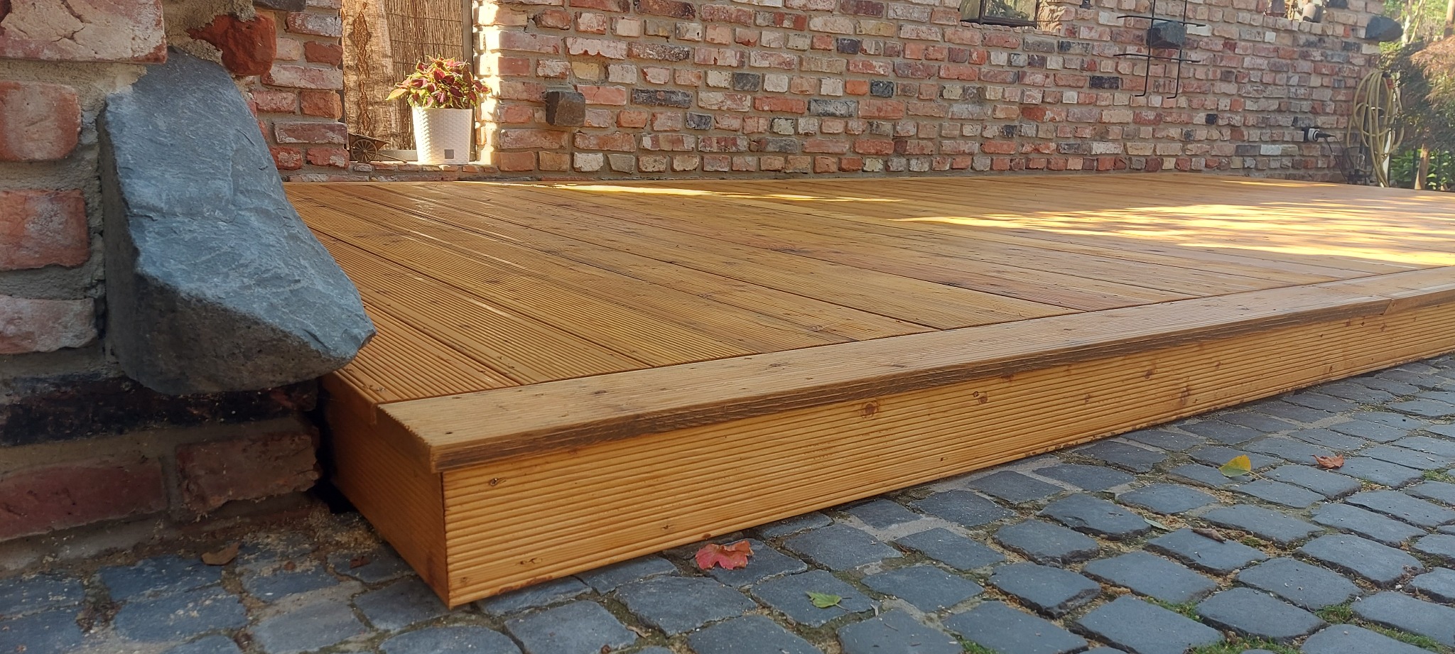 Drewniany taras z desek ryflowanych, z kamiennym akcentem przy ceglanej ścianie. W tle donica z kwiatami. Taras na kostce brukowej.