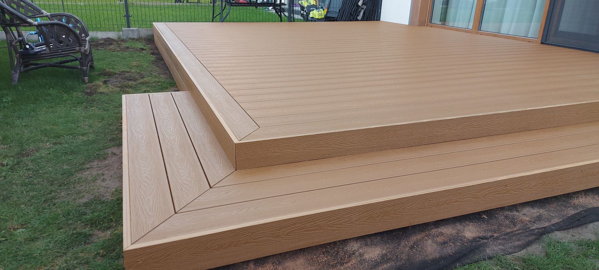 Beżowy taras z desek kompozytowych, z dwoma stopniami, prowadzący do domu. Widoczne detale wykończenia krawędzi i faktura desek.