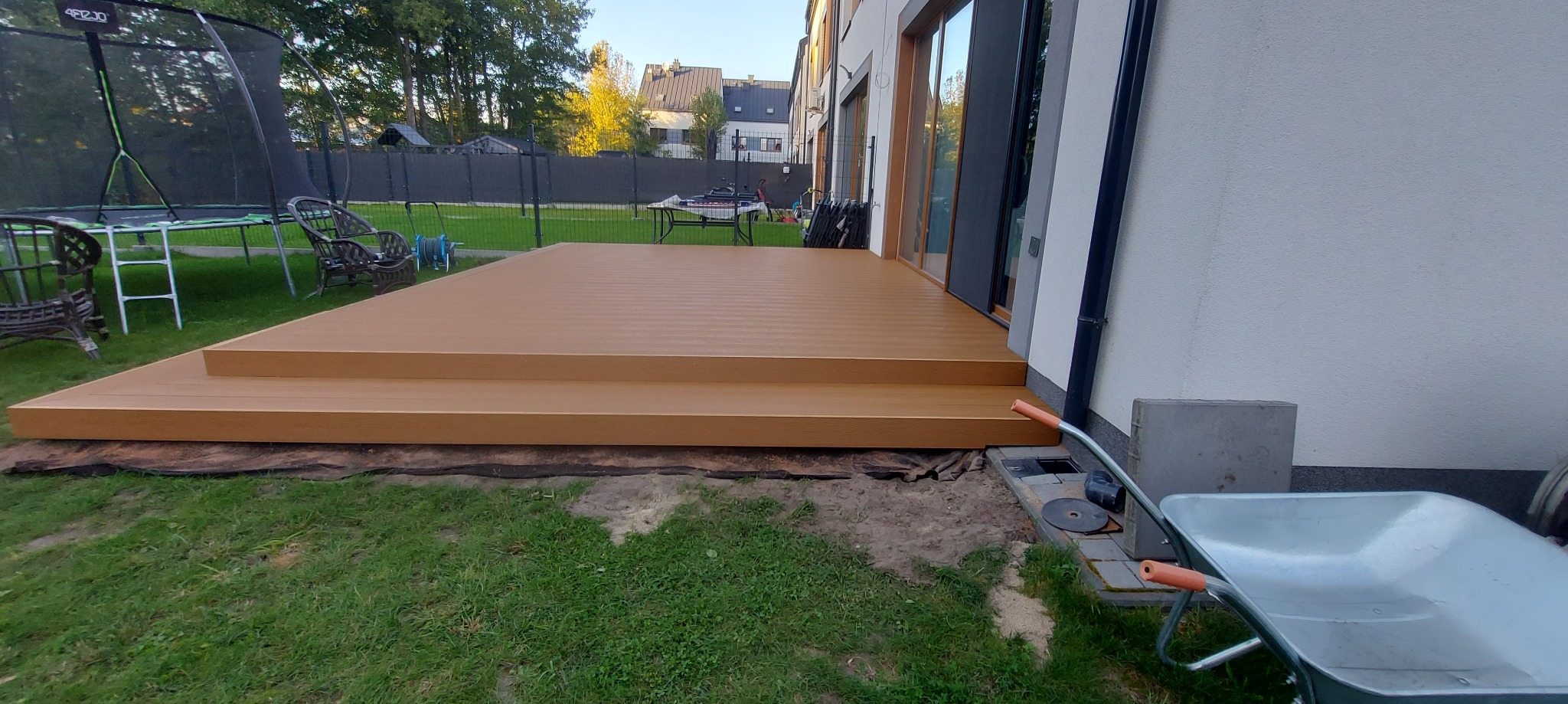 Nowoczesny taras kompozytowy z dwoma stopniami przy białym budynku. Na trawniku widoczny wózek i trampolina w tle. Słoneczny dzień, ciepłe światło.