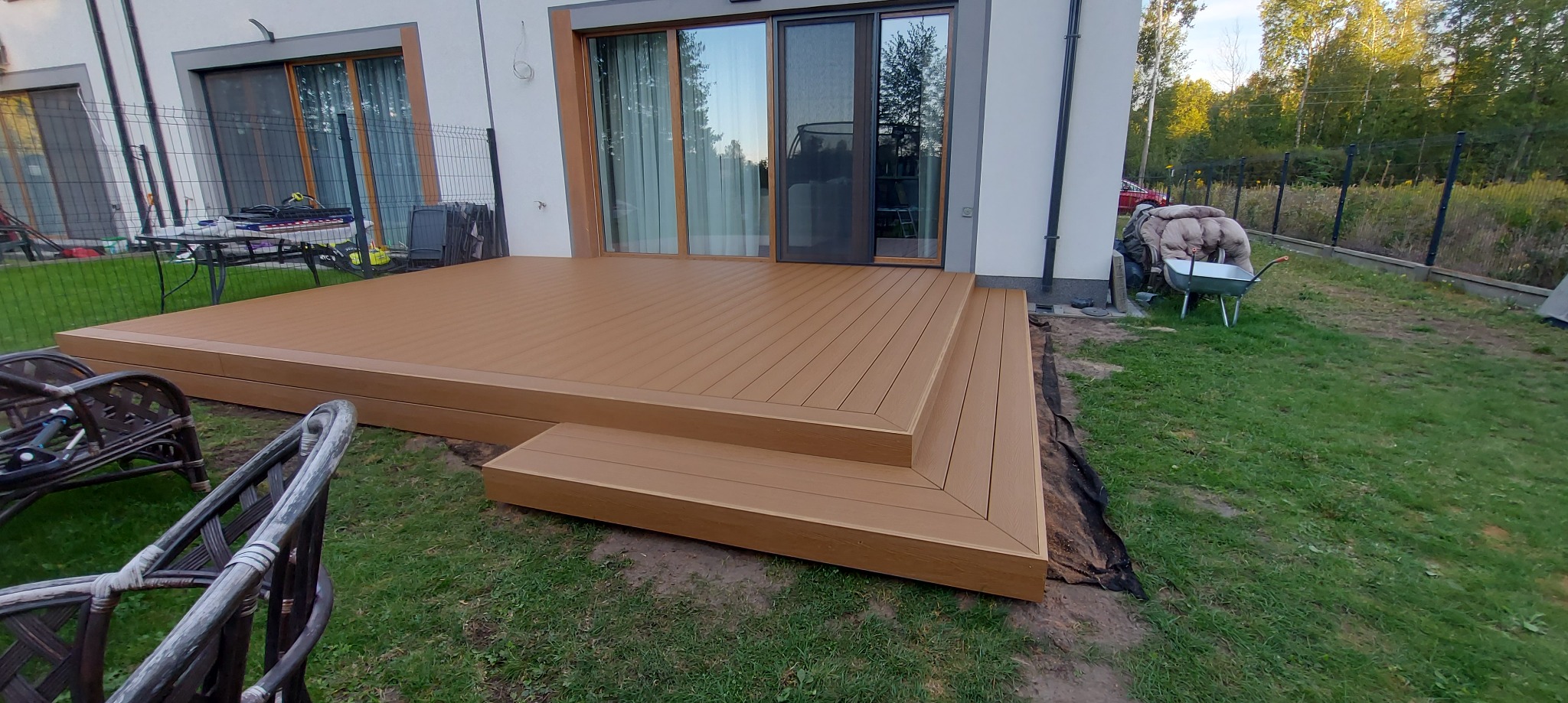 Nowoczesny taras z kompozytu w odcieniach brązu z dwoma stopniami, przylegający do białego domu z dużymi oknami. W tle ogród z wózkiem i narzędziami.