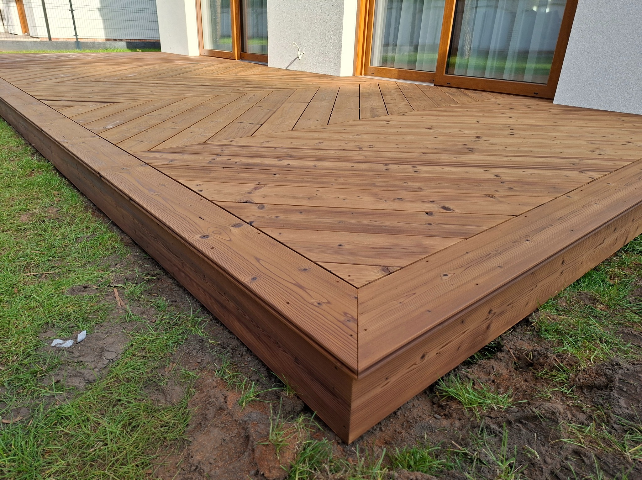 Nowoczesny taras z drewna o unikalnym wzorze ułożenia desek, przylegający do budynku z dużymi oknami, otoczony zielenią.