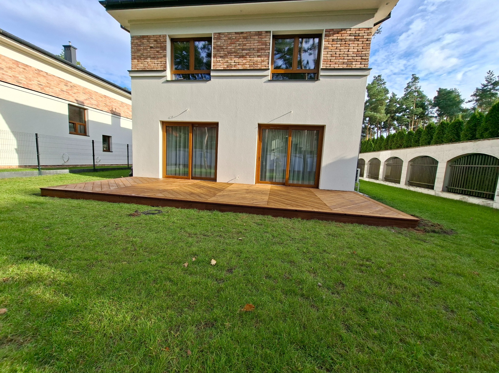 Nowoczesny taras z drewna egzotycznego przylegający do domu z jasną elewacją i ceglanymi elementami. Ułożony wzór desek w jodełkę. Soczysta zieleń trawnika.