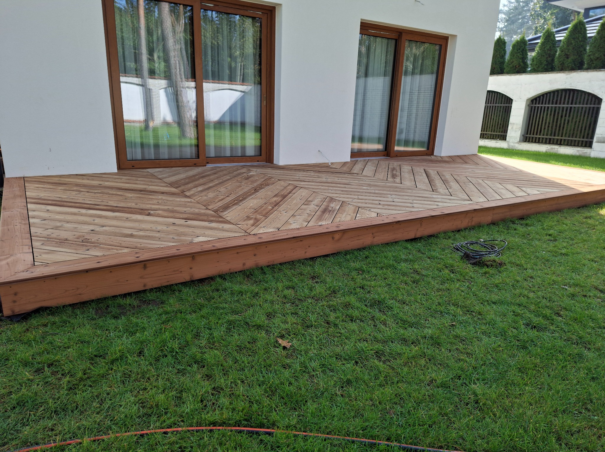 Drewniany taras z desek ułożonych we wzór jodełki, przylegający do białego budynku z brązowymi oknami, otoczony zielonym trawnikiem.