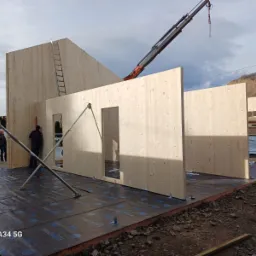 Wznoszenie ścian z drewna klejonego warstwowo na fundamencie, dźwig w tle, widoczni pracownicy i drabina oparta o ścianę szczytową.