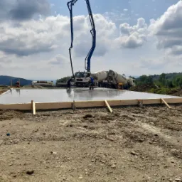 Wylewanie betonowej płyty fundamentowej z użyciem pompy do betonu i dwóch betoniarek, drewniane szalunki otaczają świeży beton, w tle wzgórza i pochmurne niebo.