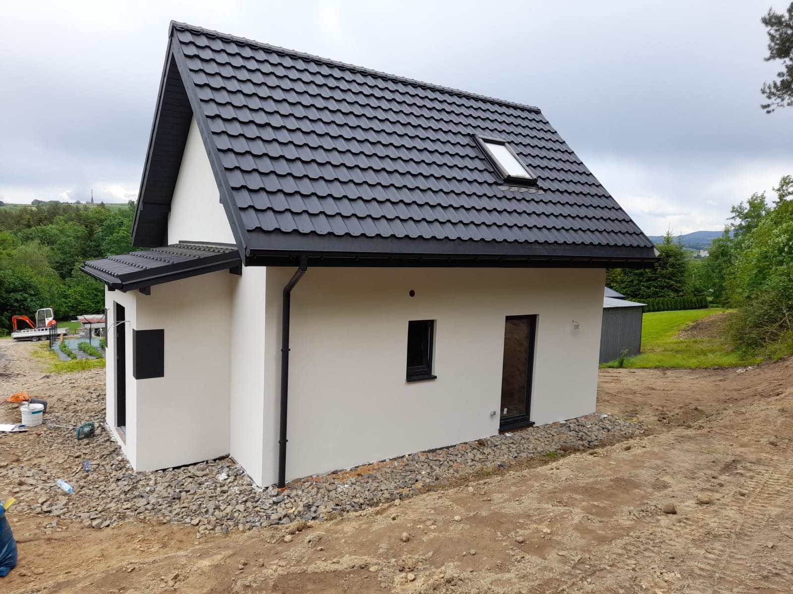 Niedawno wybudowany, parterowy dom z czarną, metalową dachówką i białymi ścianami, na jeszcze niezagospodarowanej działce z widocznymi śladami prac ziemnych.