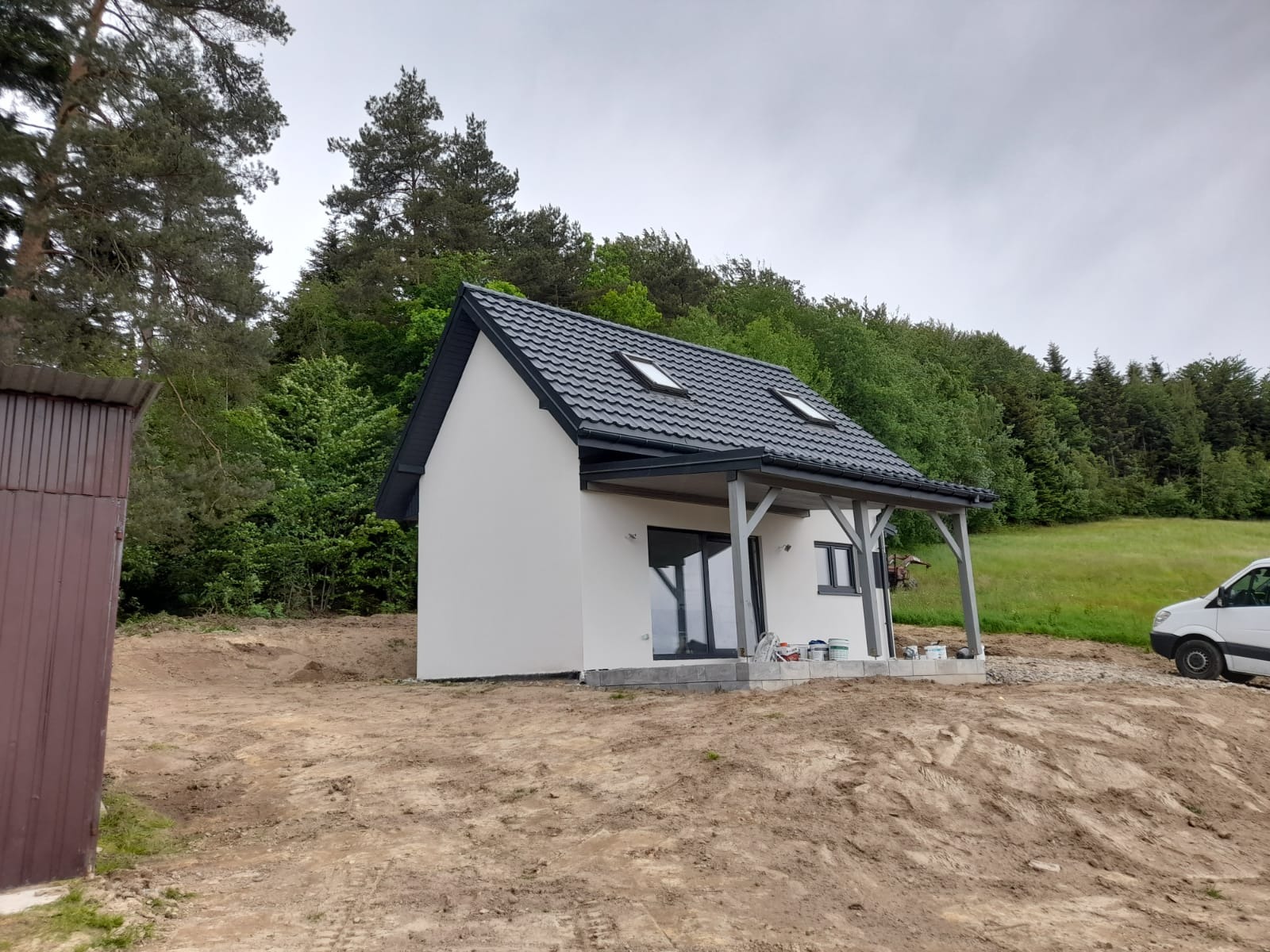 Niewielki, parterowy dom z dwuspadowym dachem krytym szarą dachówką, z drewnianą werandą i widocznymi oknami dachowymi, usytuowany na działce z widokiem na las.