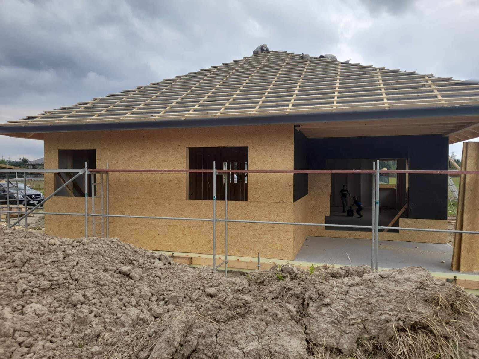 Budowa domu: widok na szkieletową konstrukcję z płyt OSB, widoczne otwory okienne i rusztowanie, w tle sterta ziemi i pracownicy na dachu.