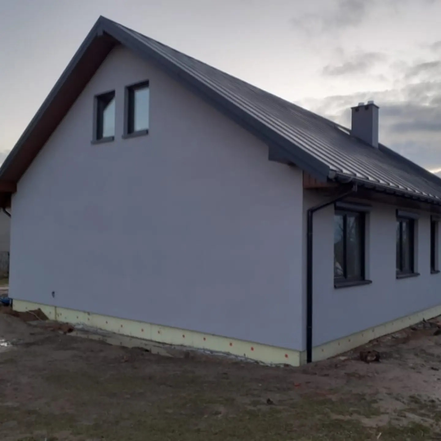 Gotowy, parterowy dom z szarą elewacją, ciemnym dachem z blachy i dwoma oknami na poddaszu, ocieplony fundament.