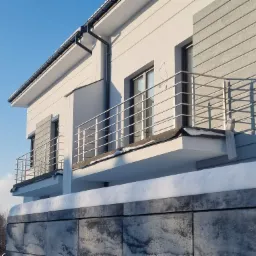 Nowoczesny dom z balkonami zabezpieczonymi metalową konstrukcją, widok z dołu na tle błękitnego nieba i ośnieżonego muru.