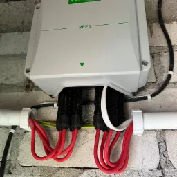 Szara skrzynka elektryczna Projoy PEFS z podłączonymi czerwonymi kablami na tle szarej, surowej ściany z cegieł.