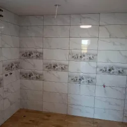 Ściany wyłożone białymi płytkami imitującymi marmur z motywem kwiatowym, drewnopodobna podłoga w narożniku pomieszczenia, widoczny fragment sufitu z lampą