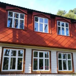 Czerwony dach z dachówki karpiówki na odnowionym, drewnianym domu z białymi oknami i ozdobną werandą, widok z dołu.