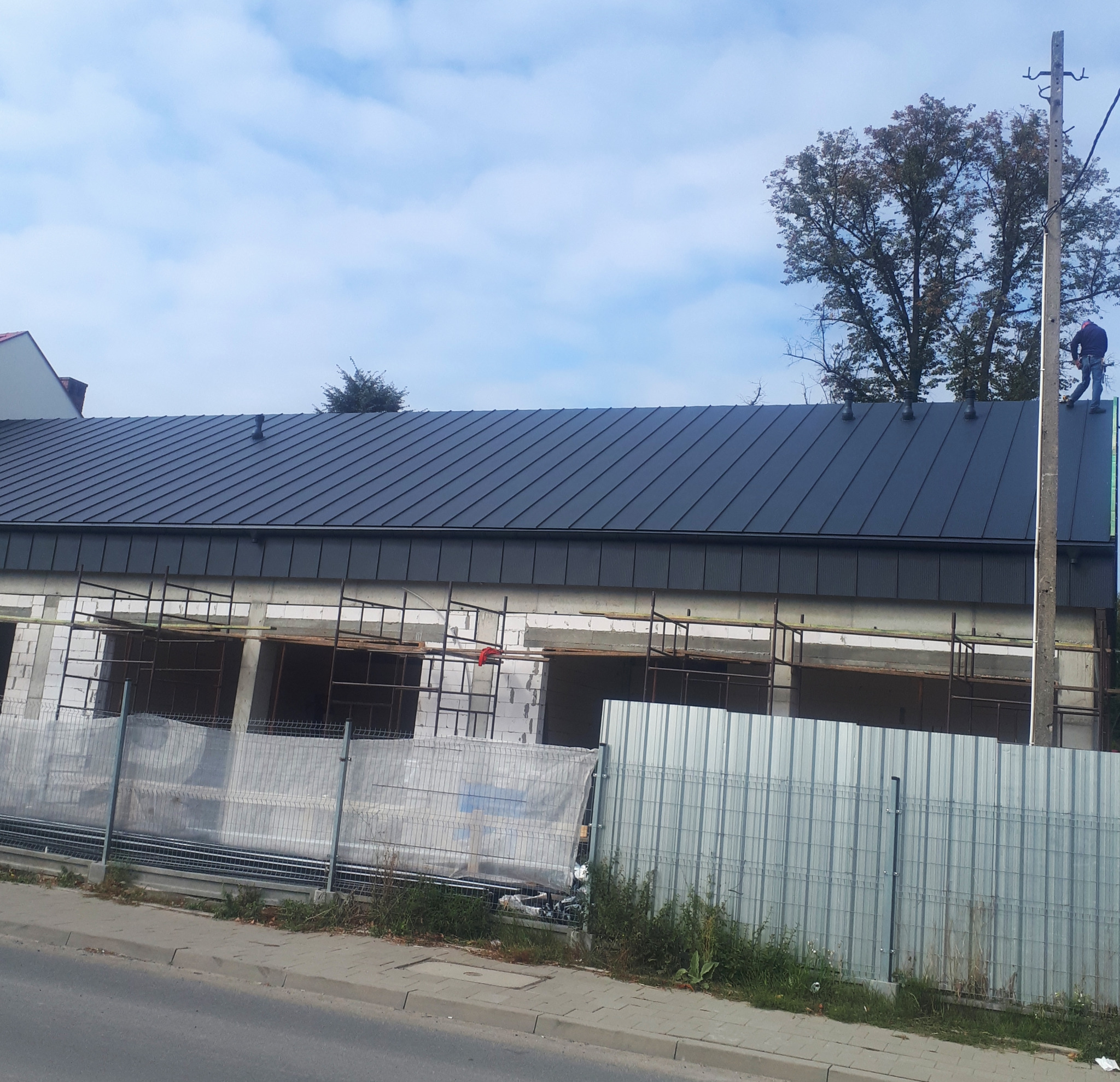 Budynek w trakcie budowy z nowym, ciemnym pokryciem dachowym. Na dachu pracuje dekarz, a na elewacji widoczne rusztowania.