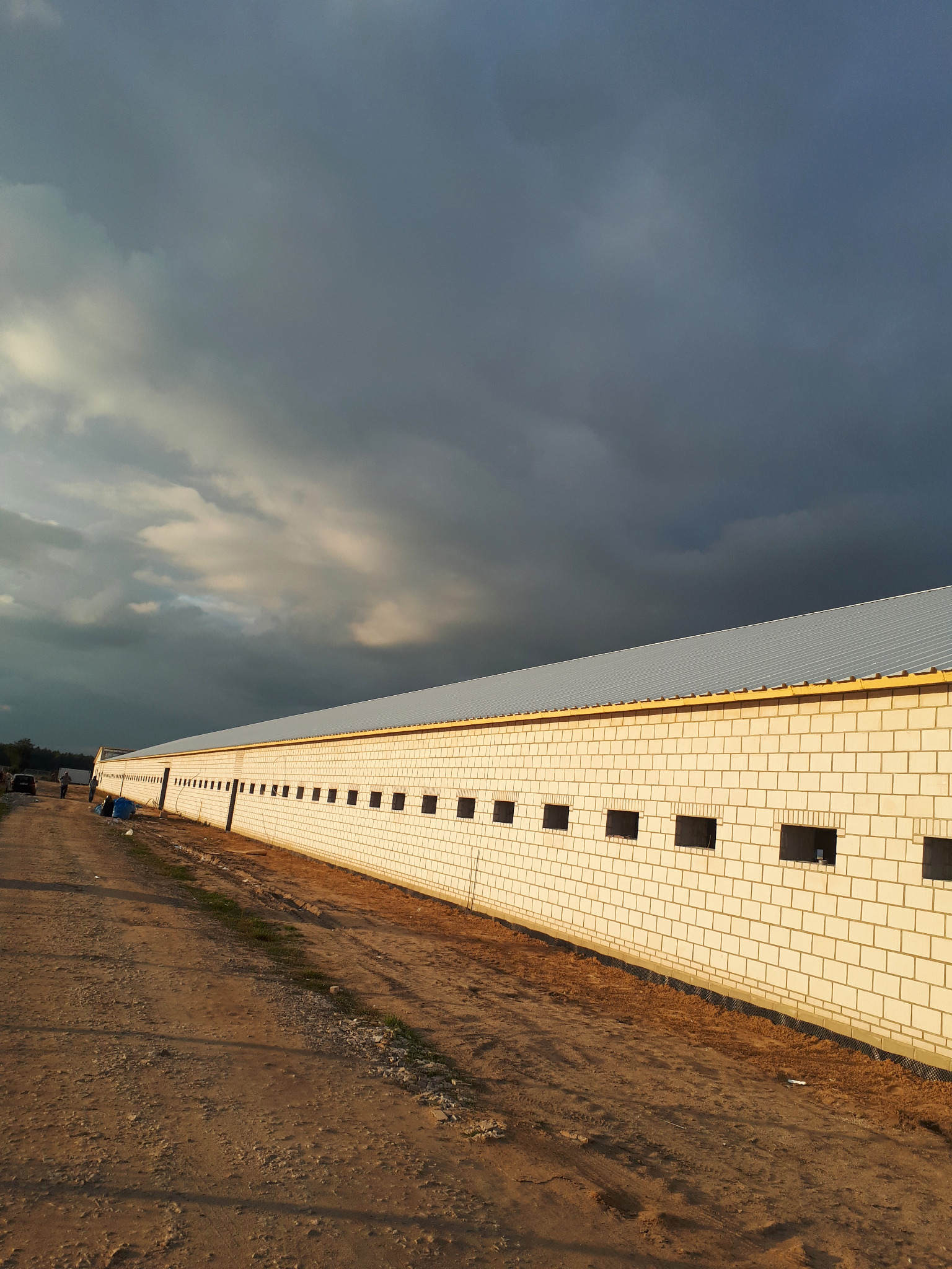 Długi budynek gospodarczy z białej cegły, z rzędem małych okien i metalowym pokryciem, sfotografowany w pochmurny dzień z perspektywy gruntu.