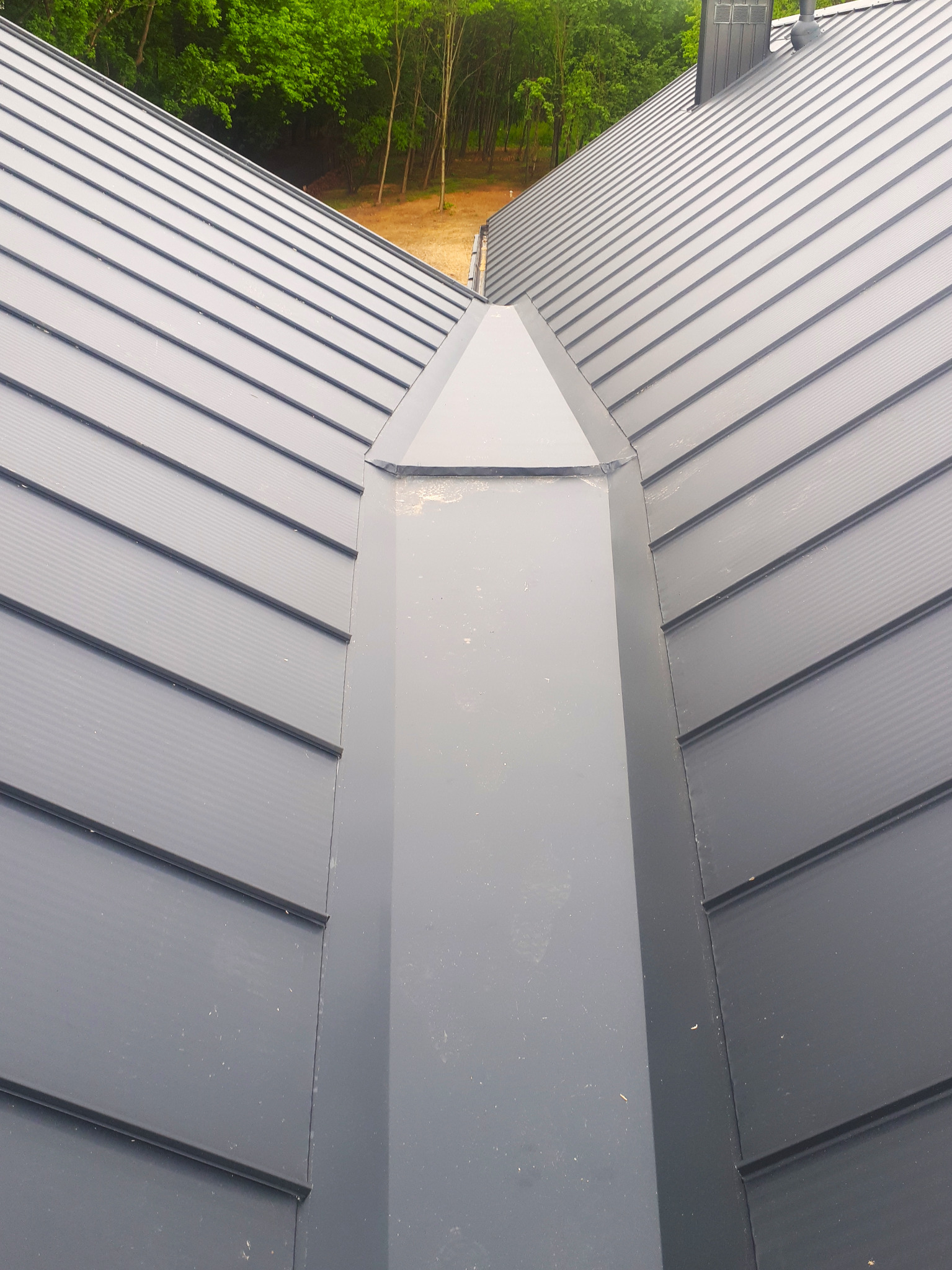 Szary dach z blachy na rąbek stojący, widok z góry na komin i połączenie połaci dachowych, w tle drzewa.