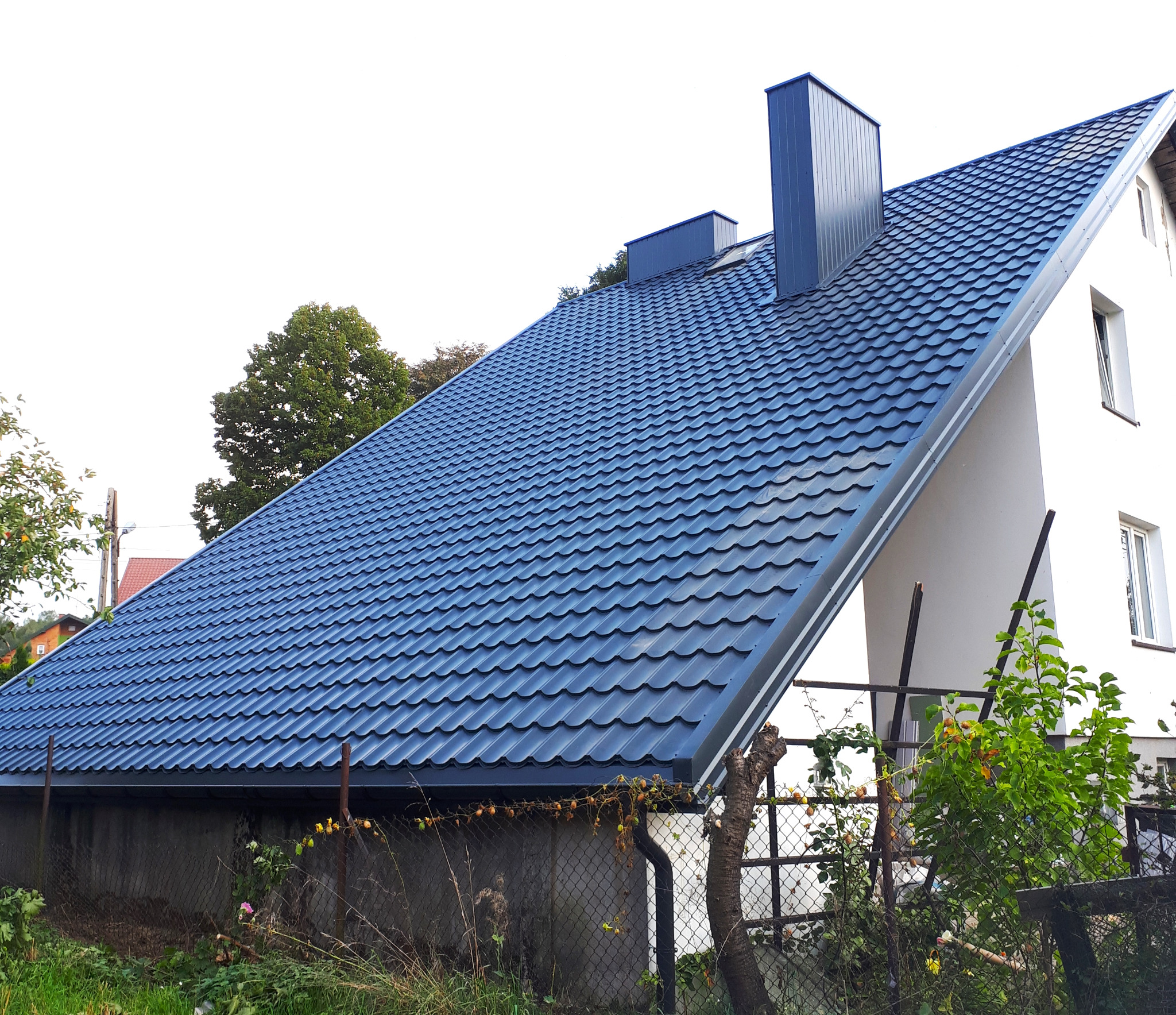 Dom jednorodzinny w Rawie Mazowieckiej z nowym, ciemnogranatowym dachem z blachodachówki i kominem w tym samym kolorze, widziany z poziomu ogrodu.
