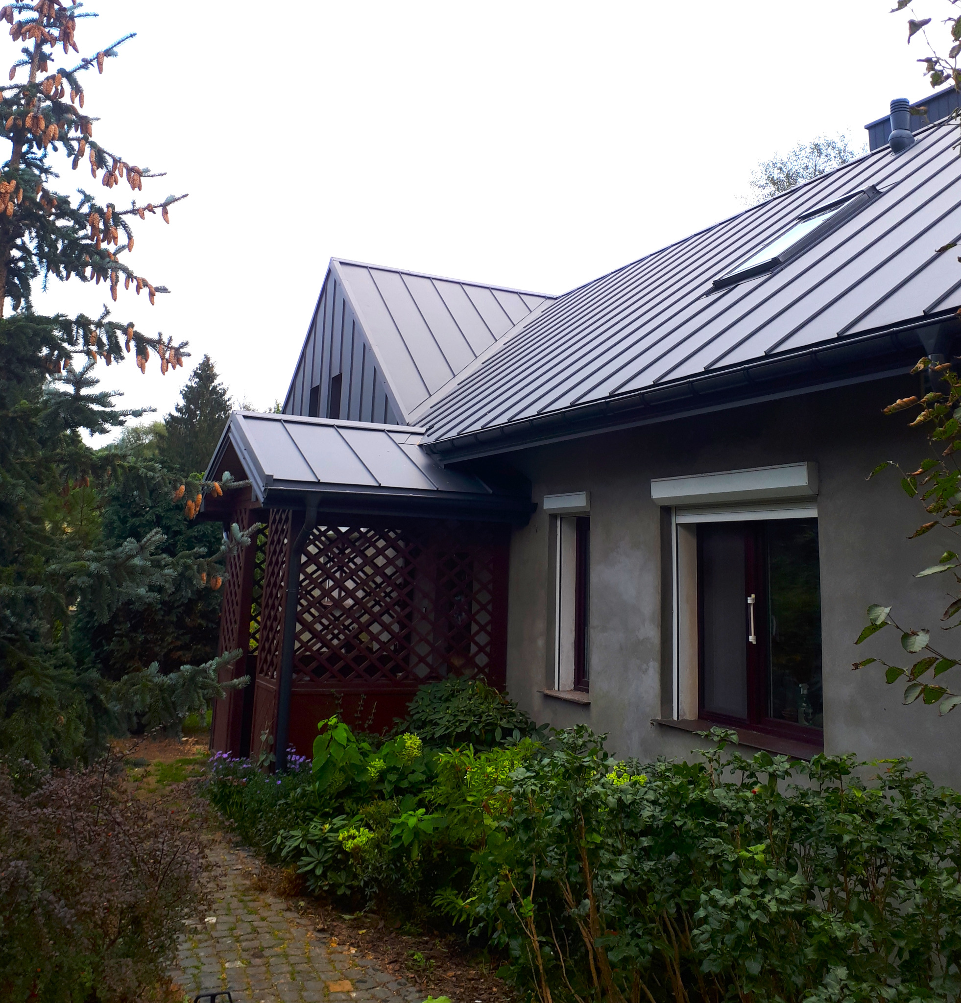 Dom jednorodzinny z nowym, ciemnoszarym dachem z blachy na rąbek stojący, widoczne okna dachowe, rynny, obróbki blacharskie i drewniana altana.