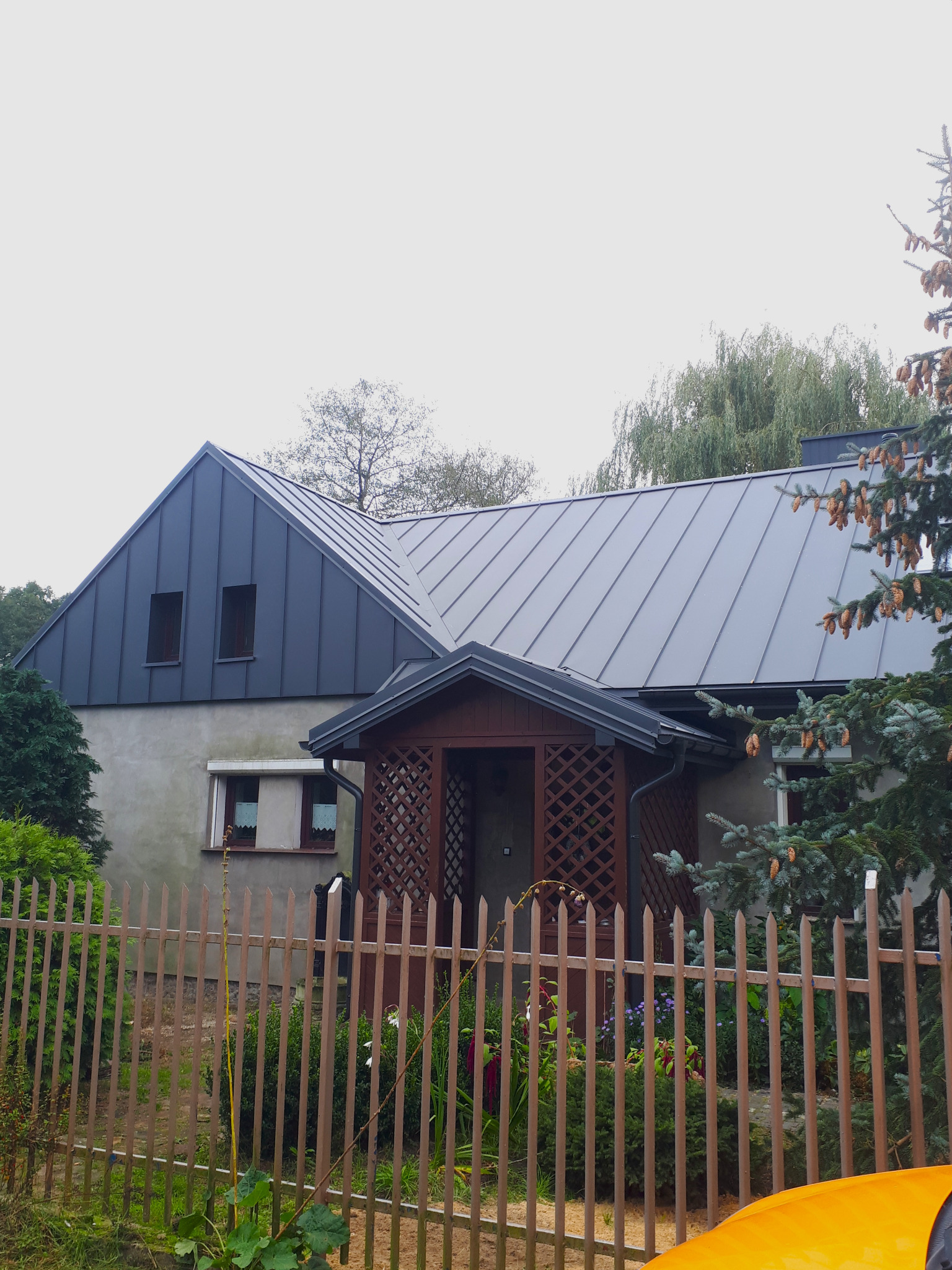 Dom jednorodzinny z nowym, szarym pokryciem dachowym z blachy na rąbek stojący, widoczne rynny i obróbki blacharskie, drewniana weranda i ogrodzenie.