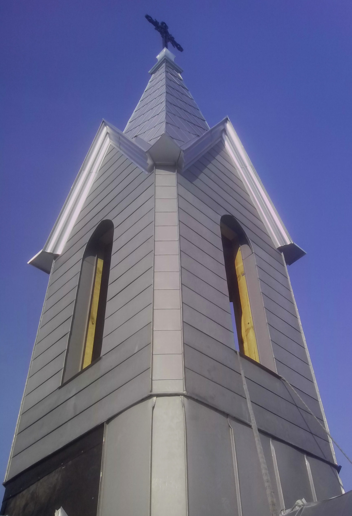 Wieża kościelna w trakcie obkładania blachą, widoczne otwarte okna i krzyż na szczycie, ujęcie z dołu na tle błękitnego nieba.