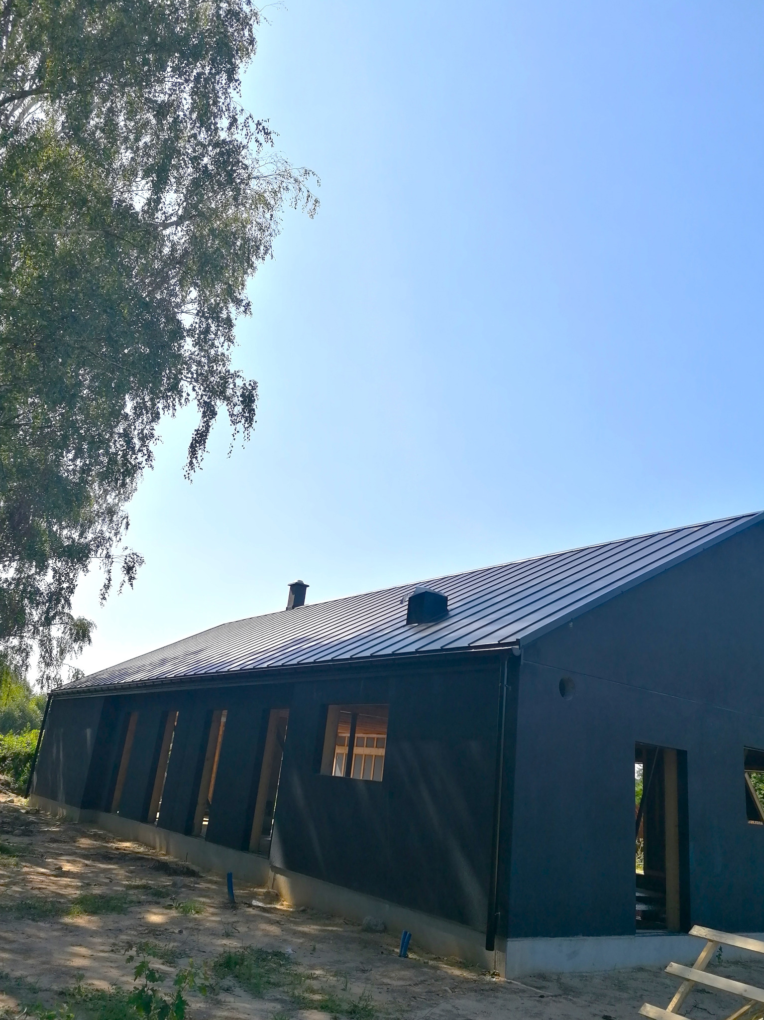 Nowoczesny dom w trakcie budowy z ciemną elewacją i metalowym pokryciem, widoczne fragmenty więźby dachowej i rynny, teren wokół w stanie surowym.