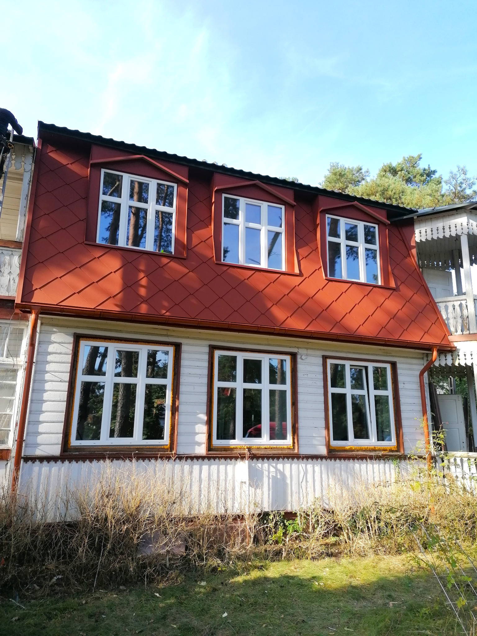 Dwupiętrowy dom z czerwonym dachem w stylu rustykalnym, widok z dołu, okna z białymi ramami, elewacja drewniana, częściowo zasłonięta przez wysokie trawy.