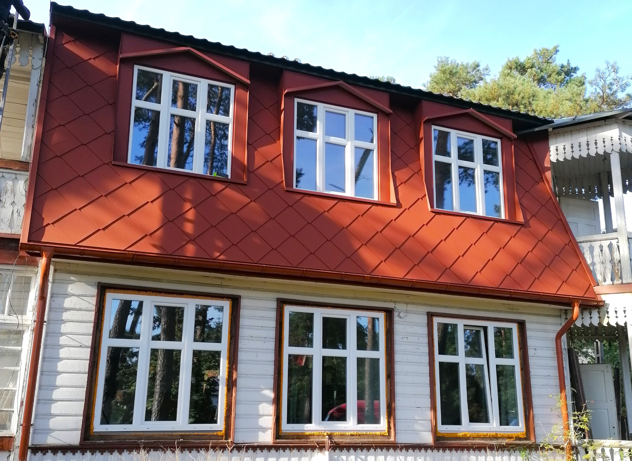 Czerwony dach z dachówki karpiówki na odnowionym, drewnianym domu z białymi oknami i ozdobną werandą, widok z dołu.