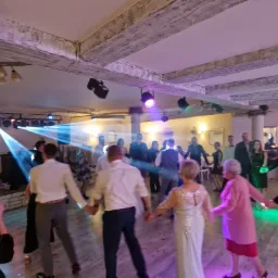 Uczestnicy wesela tańczą w sali z dekoracyjnymi belkami stropowymi i oświetleniem scenicznym, w tle widoczny parkiet i goście.