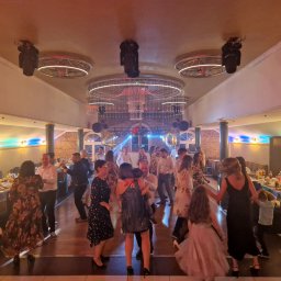 A & G Arkadiusz Szymański - Sala balowa z parkietem, oświetlona dekoracyjnymi lampami sufitowymi, w której tańczy grupa ludzi w strojach wieczorowych, z widocznymi nakrytymi stołami w tle.