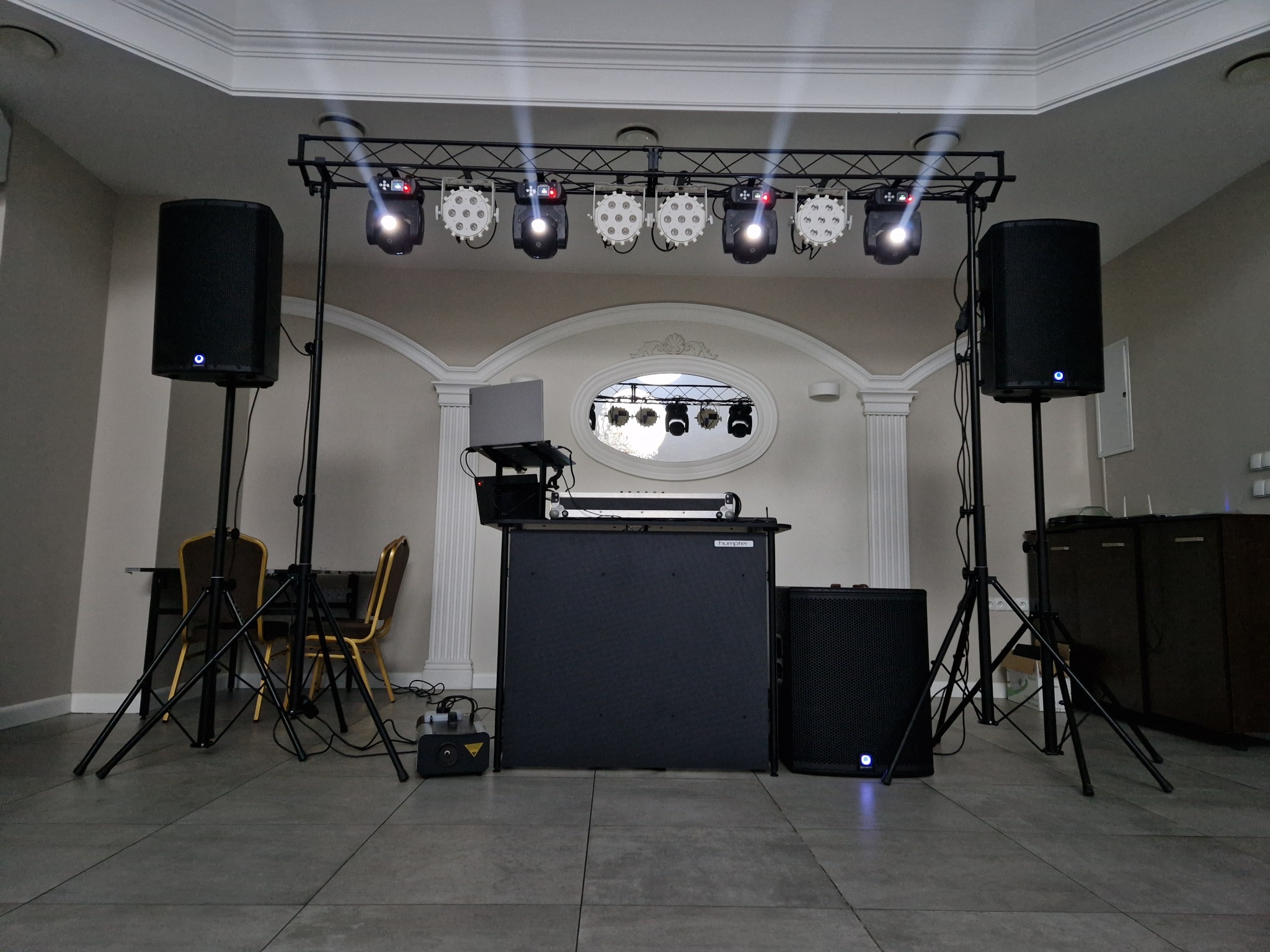 Rozstawiony sprzęt DJski z laptopem, mikserem, głośnikami na statywach i oświetleniem dyskotekowym w sali z kolumnami i dekoracyjnym oknem