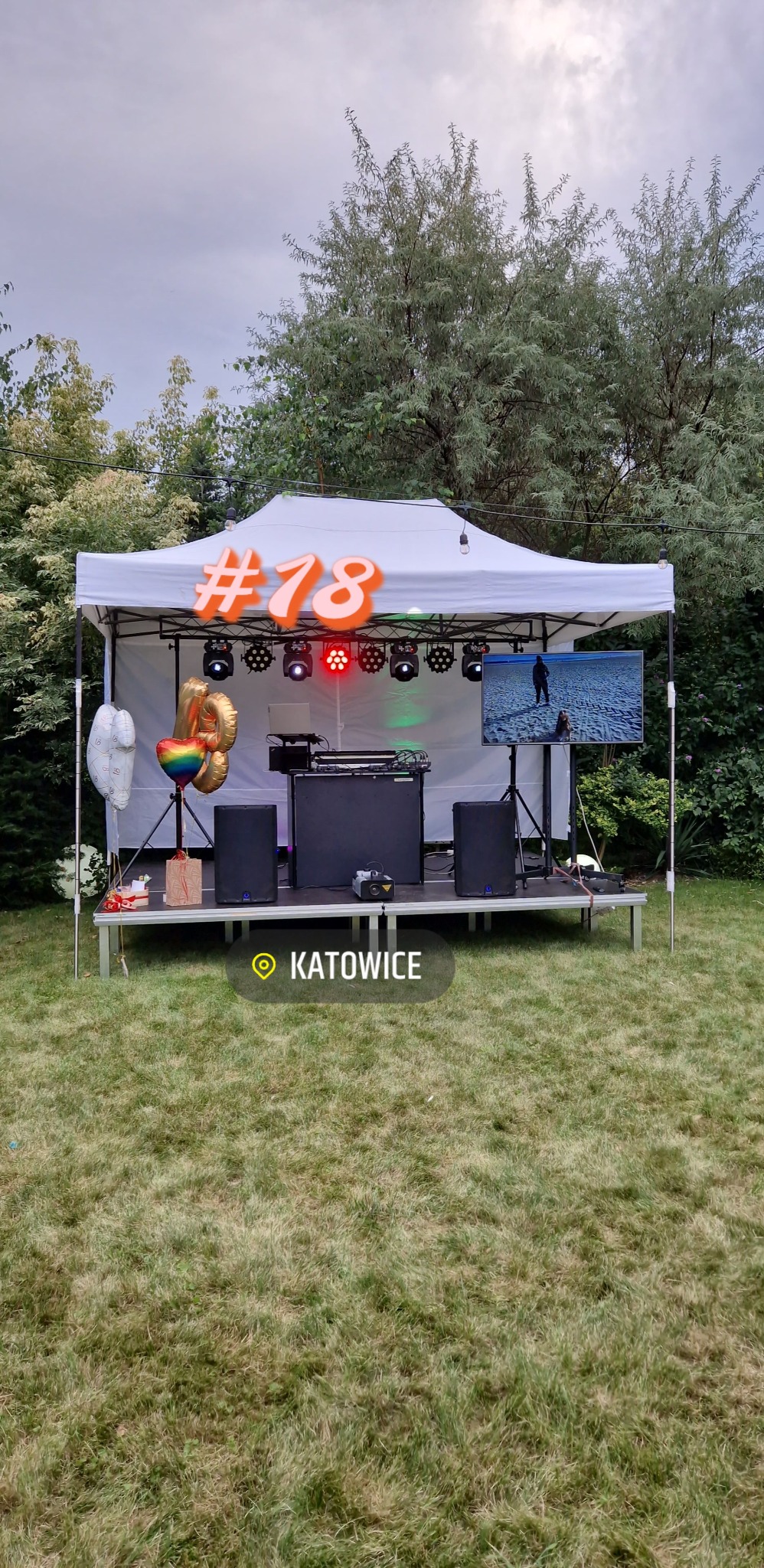 Scena plenerowa pod białym namiotem, przygotowana do imprezy osiemnastkowej, z nagłośnieniem, oświetleniem, ekranem wyświetlającym plażę i balonami w kształcie cyfr.