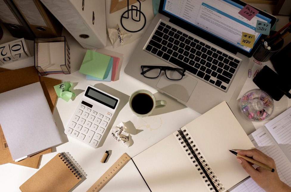 Widok z góry na biurko z laptopem, kalkulatorem, notatnikiem, kawą i dłonią piszącą, otoczone dokumentami i biurowymi akcesoriami, sugerujące pracę biurową.