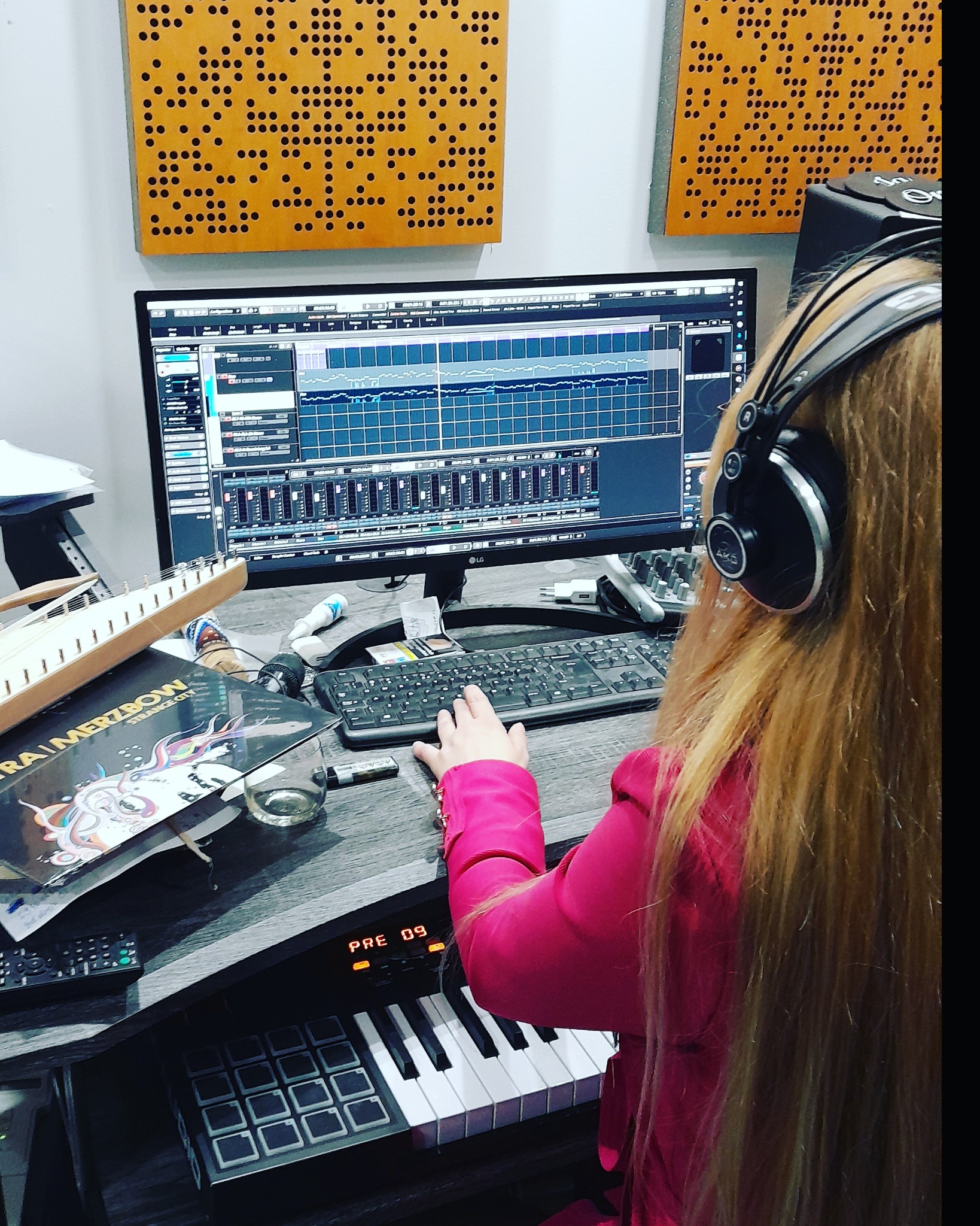 Kobieta w słuchawkach edytuje dźwięk na monitorze komputera, widoczne panele akustyczne na ścianie, kontroler MIDI i płyta winylowa na biurku.